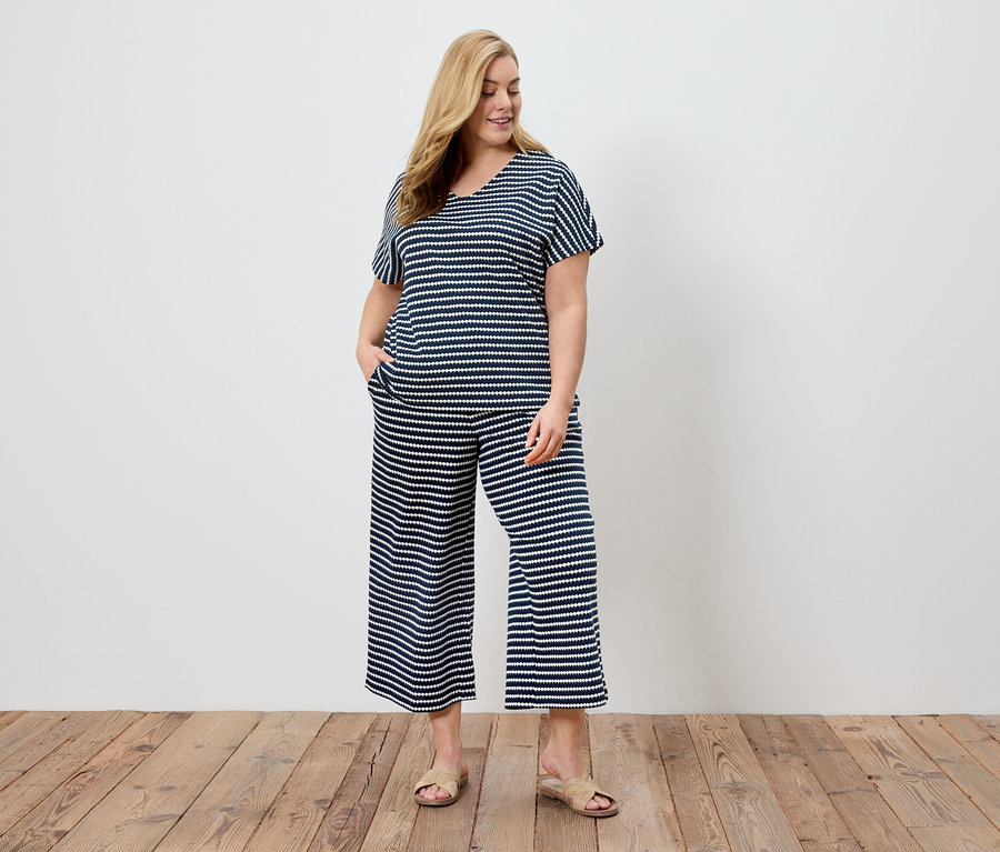 Frau mit blonden Haaren trägt einen gestreiften marineblauen und weißen Pyjama und Sandalen vor einer weißen Wand.