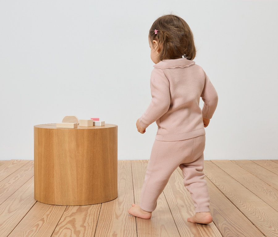 Ein Kleinkind in rosa Strickkleidung geht barfuß auf einem Holzboden zu einem runden Holztisch mit Holzspielzeug.