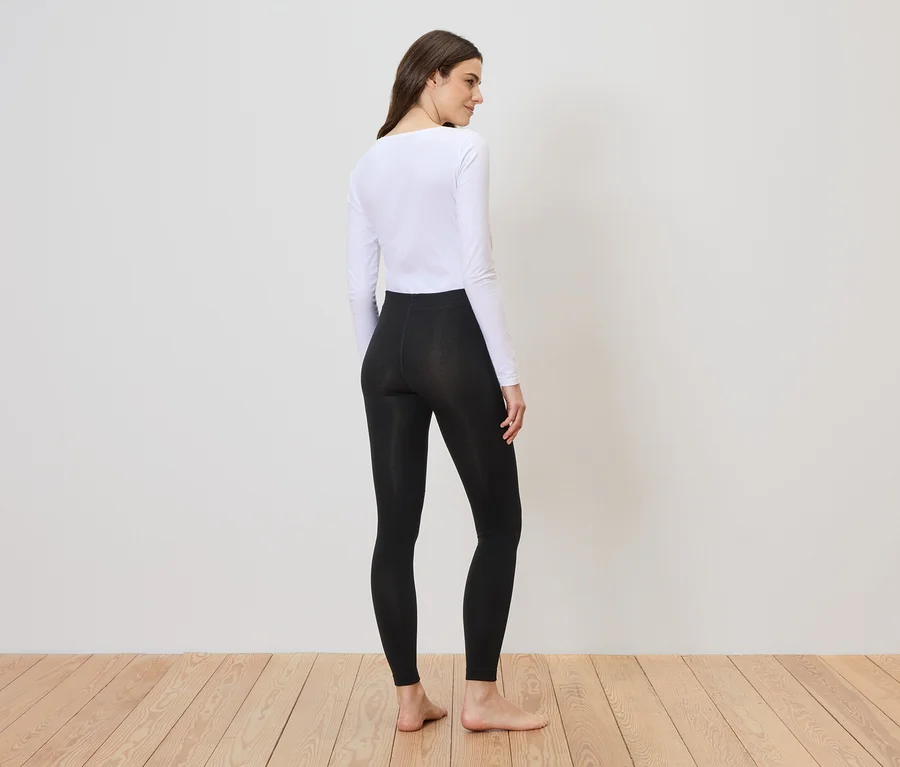 Frau mit weißem Oberteil und schwarzer Leggings steht auf Holzboden vor einer weißen Wand und blickt über ihre Schulter.