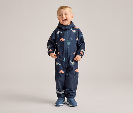 Ein lachender Junge steht in einem marineblauen Overall mit Dinosaurier-Muster und blauen Schuhen vor einem hellen Hintergrund.