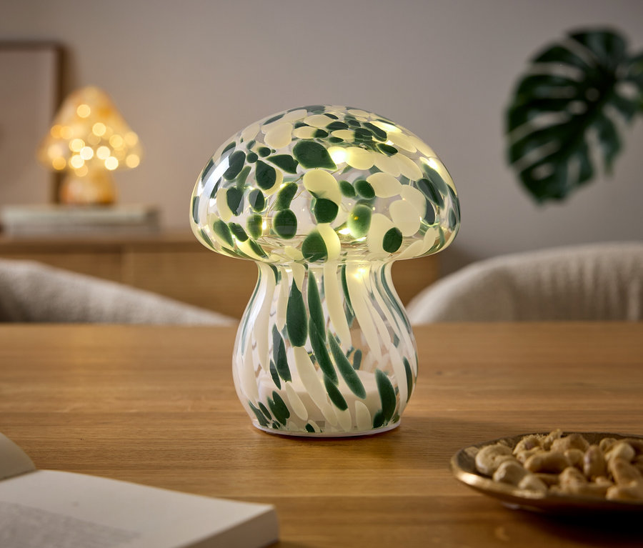 Pilzförmige Lampe mit grün-weißem Muster und Lichtern steht auf einem Holztisch. Daneben liegen ein offenes Buch und eine Schale mit Cashewkernen. Im Hintergrund ist eine weitere Pilzlampe und eine Pflanze zu sehen.