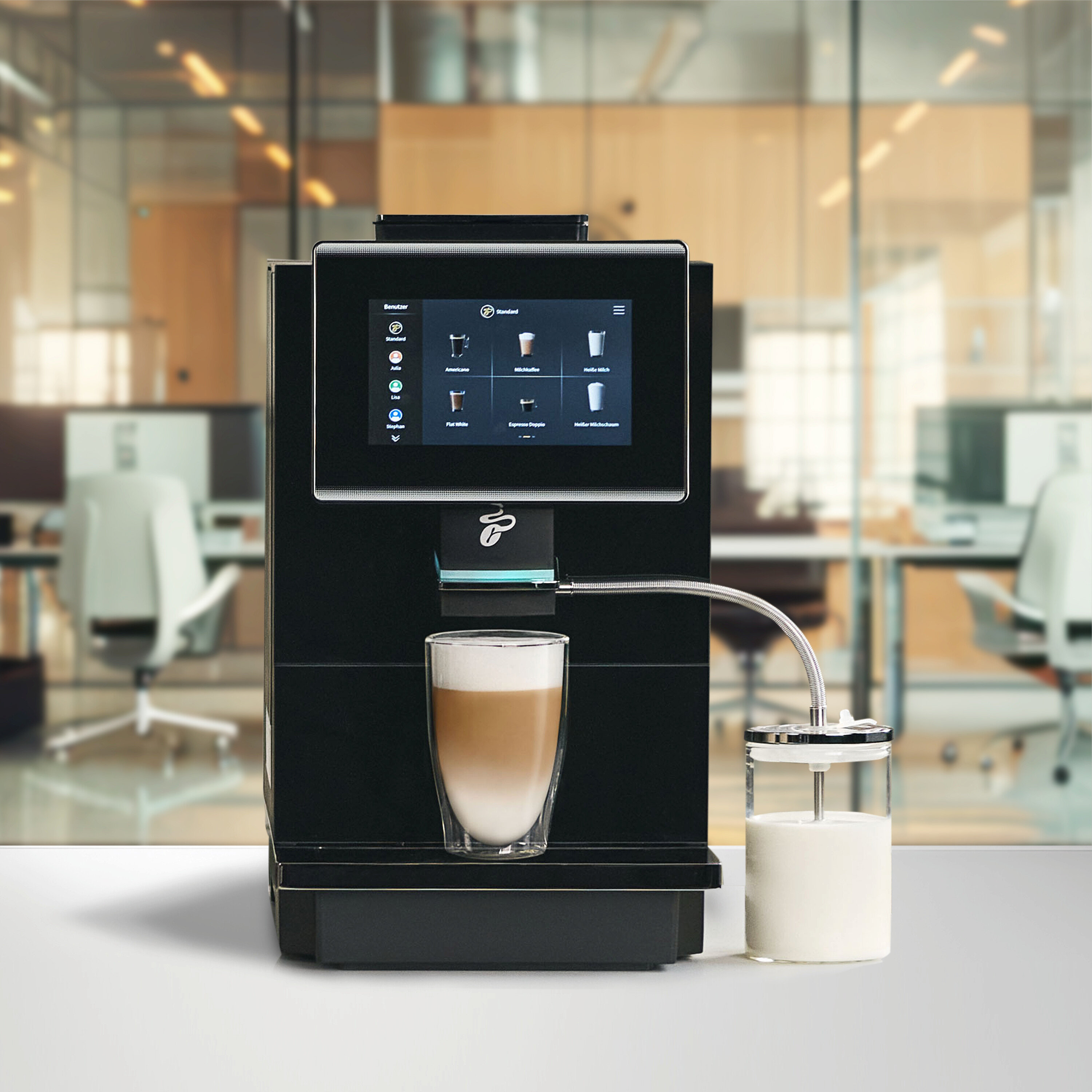 Schwarze Kaffeemaschine mit Touchscreen-Display, das Kaffeeoptionen anzeigt, einem Glas Latte Macchiato und einem Milchbehälter, der über einen Schlauch verbunden ist, in einer Büroumgebung.