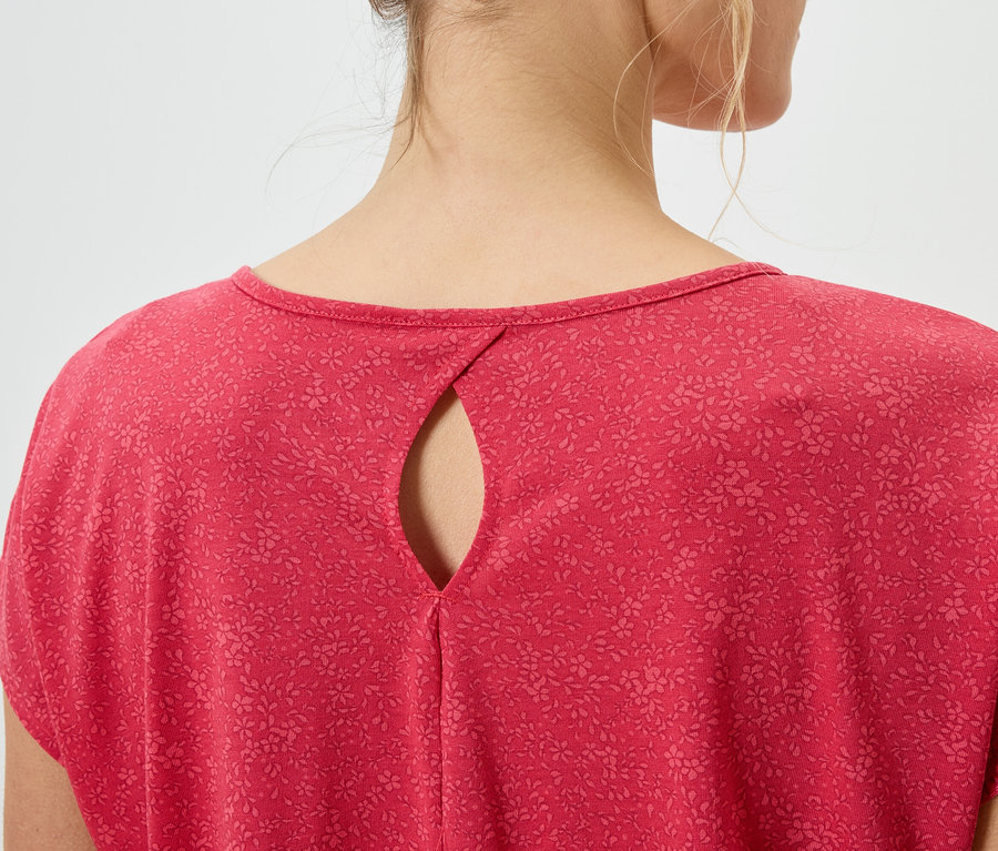 Rückansicht einer Frau mit blonden Haaren, die ein rotes, kurzärmeliges Oberteil mit Blumenmuster und einem tropfenförmigen Ausschnitt trägt.