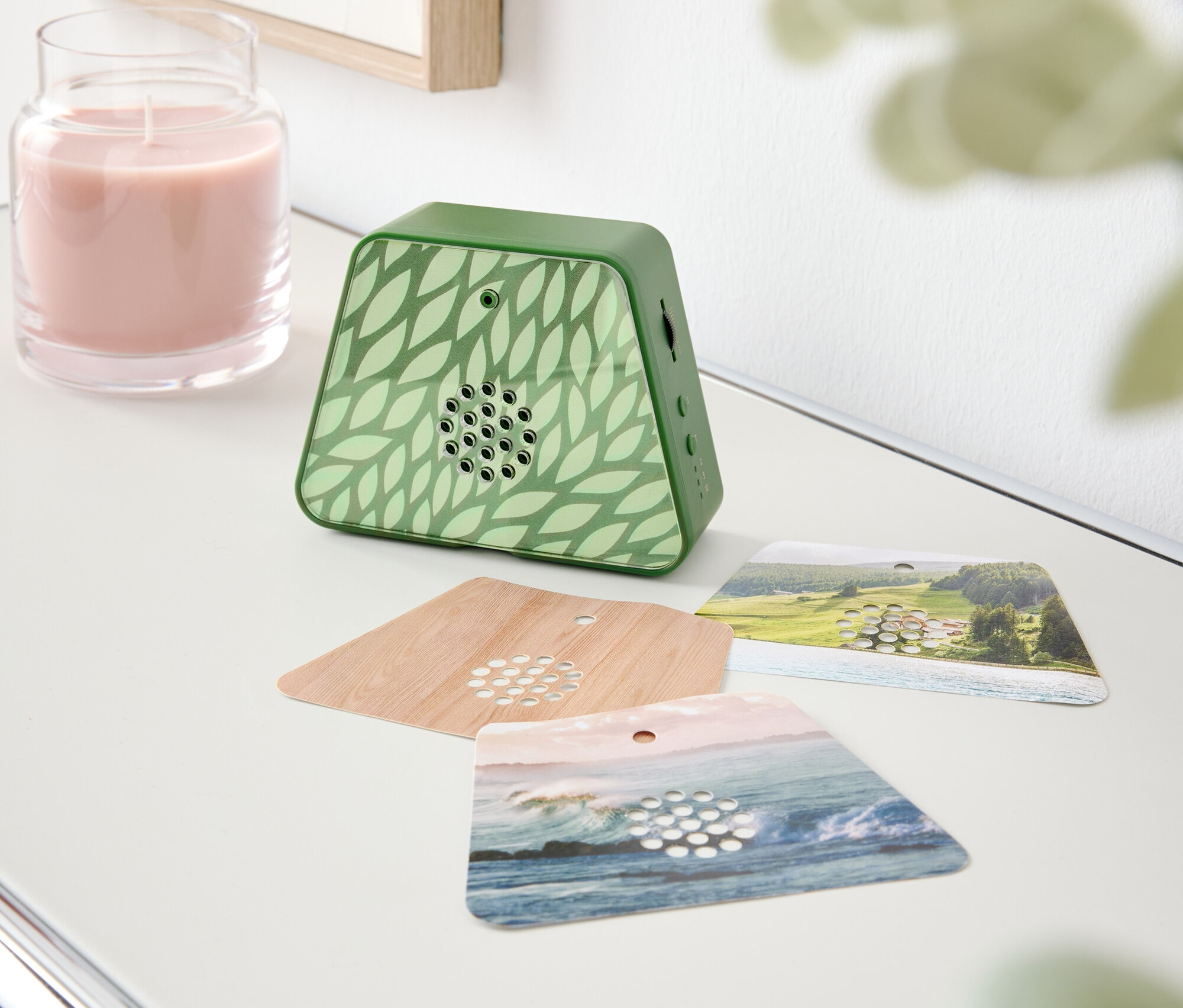 Auf einem Tisch stehen eine rosa Kerze, ein grüner Duftspender mit Blattmuster und drei Duftkarten.