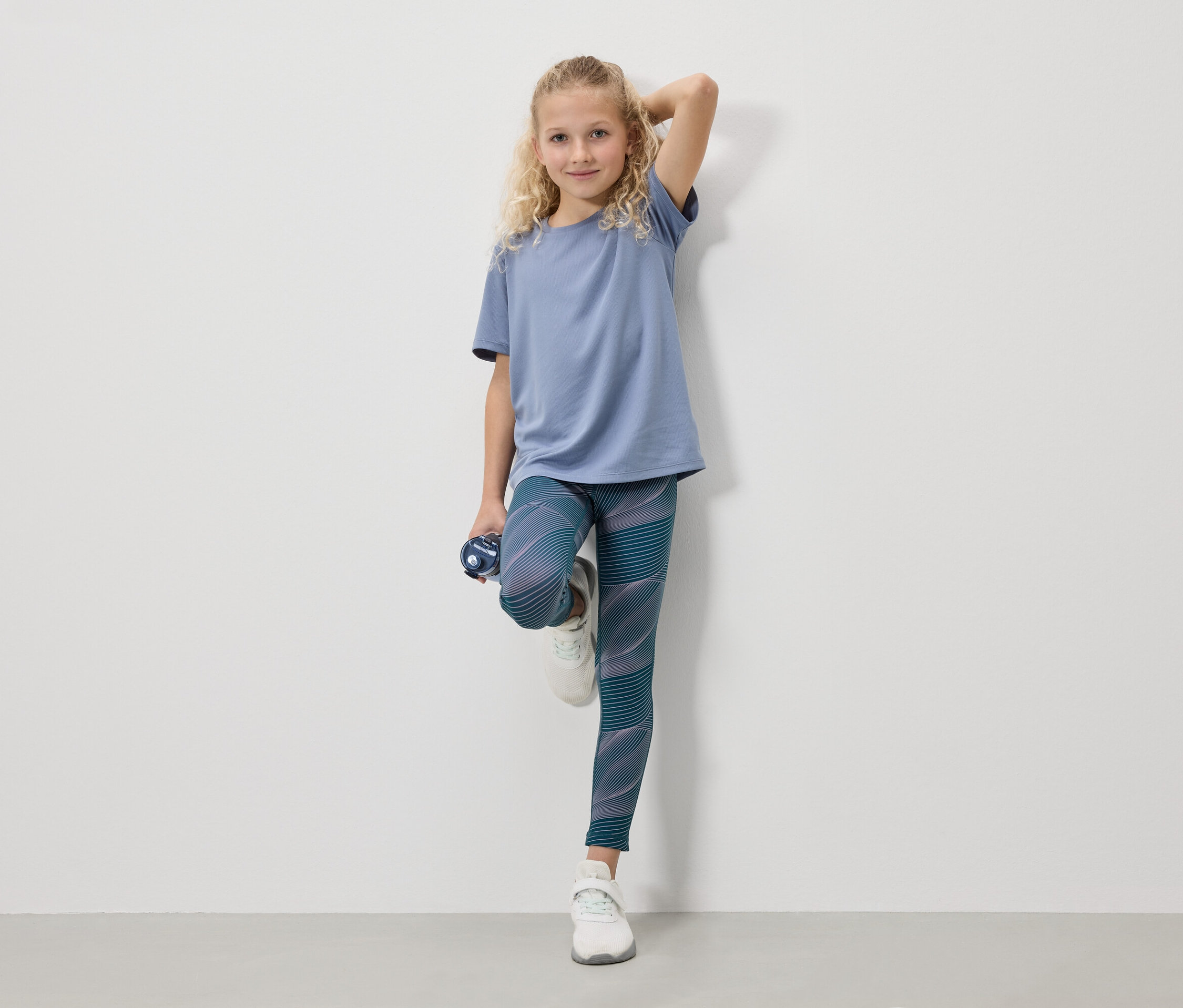 Ein blondes Mädchen in einem blauen T-Shirt, Leggings und weißen Turnschuhen steht mit einer Wasserflasche vor einer weißen Wand.