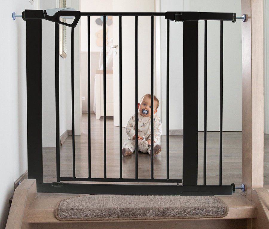 Ein Baby sitzt hinter einem schwarzen roba easySafe+ Tür-und-Treppen-Schutzgitter, das oben an einer Treppe installiert ist. Das Baby trägt einen Strampler und hat einen blauen Schnuller im Mund.