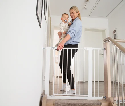 Eine Frau hält ein Baby und öffnet ein weißes roba easySafe+ Tür- und Treppen-Schutzgitter am oberen Ende einer Treppe.