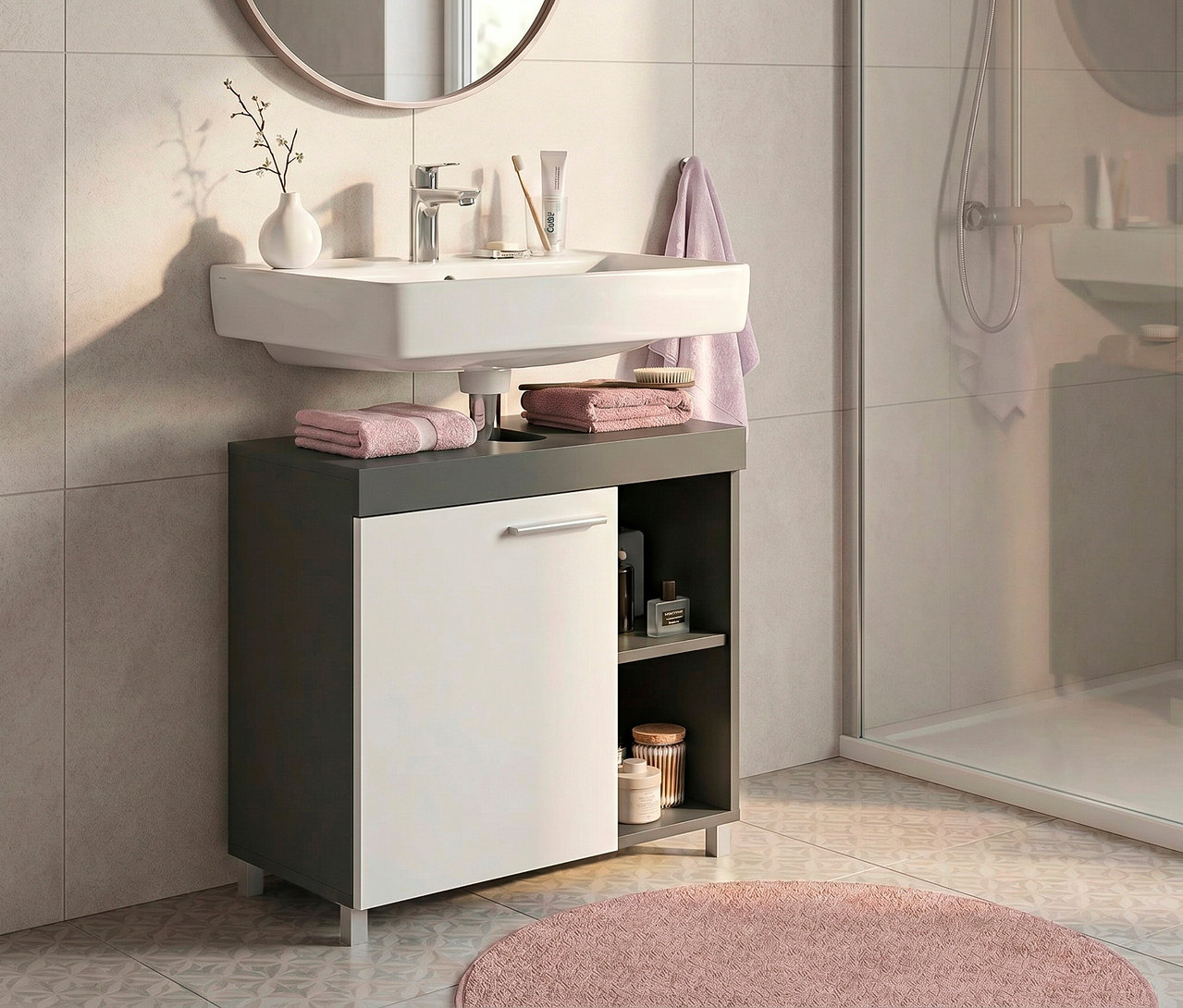 Modernes Badezimmer mit weißem Waschbecken über einem grau-weißen Waschtischunterschrank. Über dem Waschbecken hängt ein runder Spiegel mit roségoldenem Rahmen. Rosa Handtücher liegen auf dem Waschtisch, eine runde rosa Badematte auf dem gemusterten Boden. Eine Glasdusche ist rechts sichtbar.