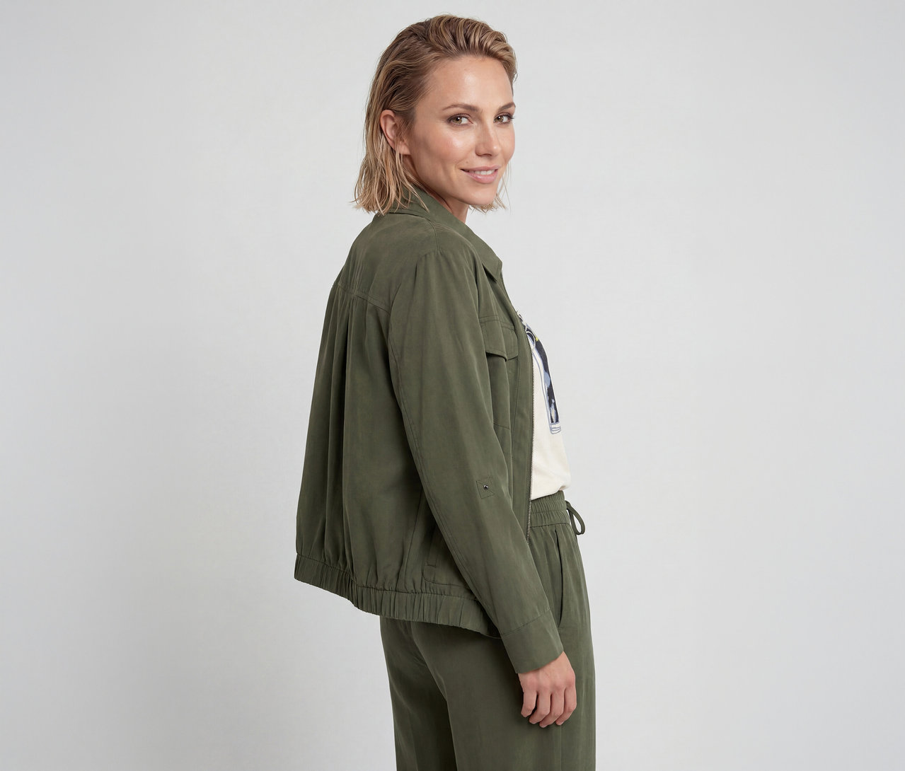Frau, leicht gedreht, blickt über ihre Schulter, trägt eine olivgrüne Jacke mit hochgekrempelten Ärmeln, ein weißes Grafik-T-Shirt und passende olivgrüne Hose.