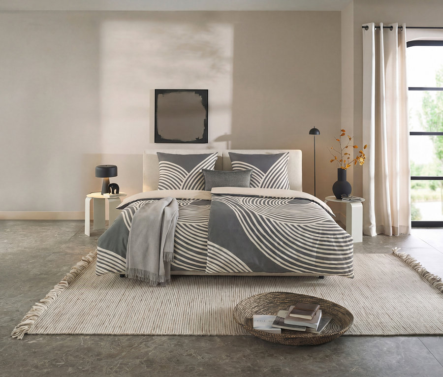 Modernes Schlafzimmer mit einem Bett, das mit der SCHÖNER WOHNEN – Kollektion Bettwäsche in Grau mit weißem Wellenmuster bezogen ist. Helle Nachttische, ein Teppich und ein Korb mit Büchern ergänzen den Raum.
