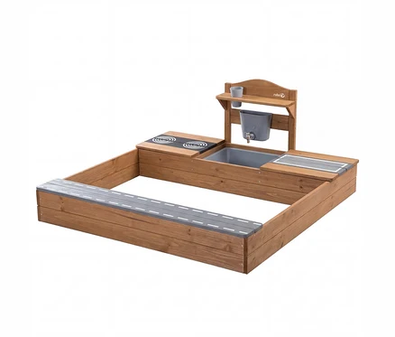 Rechteckiger Sandkasten aus Holz mit integrierter Matschküche. Die Küche hat eine Herdplatte, ein graues Waschbecken, einen Wasserhahn mit Eimer und ein Regal mit zwei Tassen. Eine Seite des Sandkastens dient als Sitzbank mit grauer Fahrbahnmarkierung.