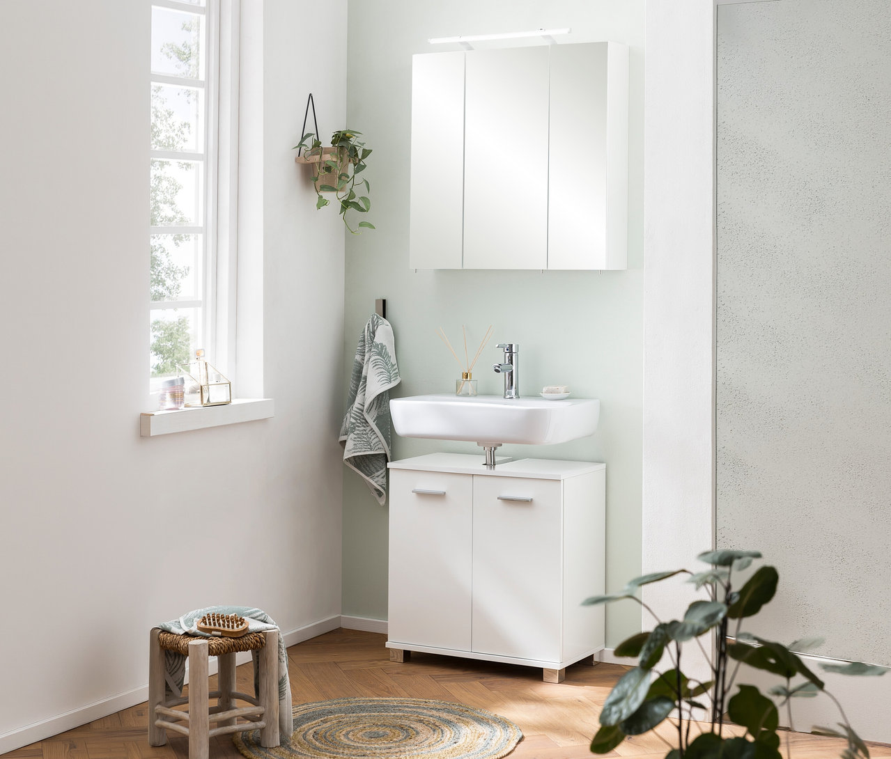 Modernes Badezimmer mit hellgrüner Wand, weißem Spiegelschrank »Dorina« mit Licht, 60 cm, über einem Waschbecken mit Unterschrank, daneben ein Fenster und ein Hocker.