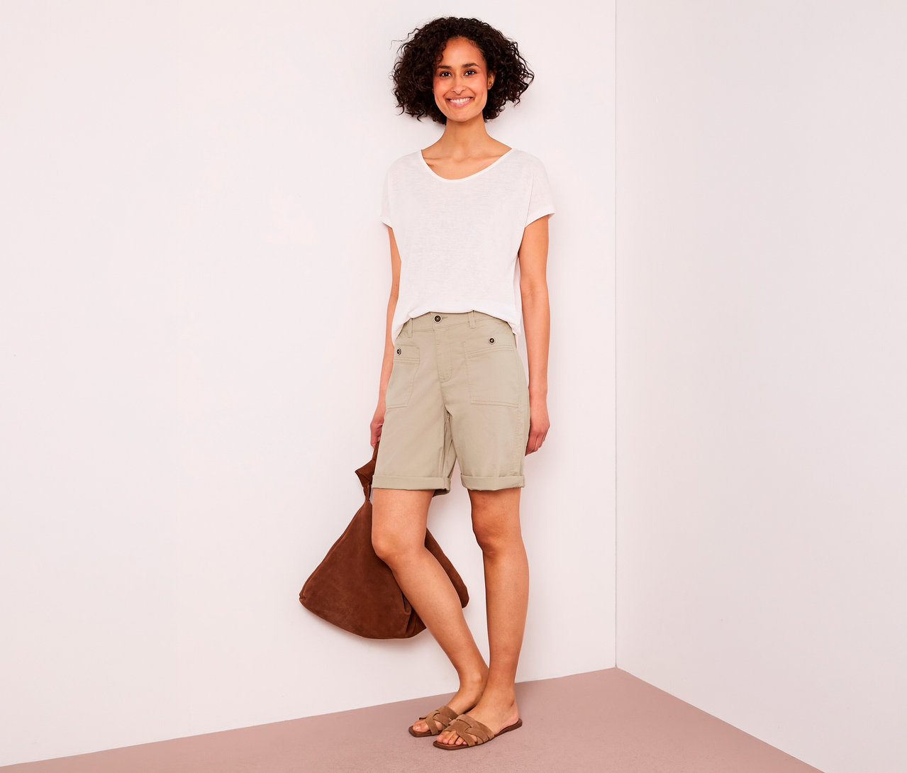 Lachende Frau mit kurzen Locken trägt ein weißes T-Shirt, beigefarbene Street One Bermuda „Yulius“ und braune Sandalen, eine braune Tasche haltend.