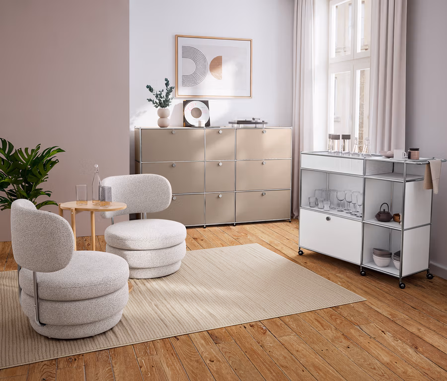Modernes Wohnzimmer mit zwei hellgrauen Sesseln, einem runden Anstelltisch aus Massivholz, beigem Sideboard und weißem Servierwagen.