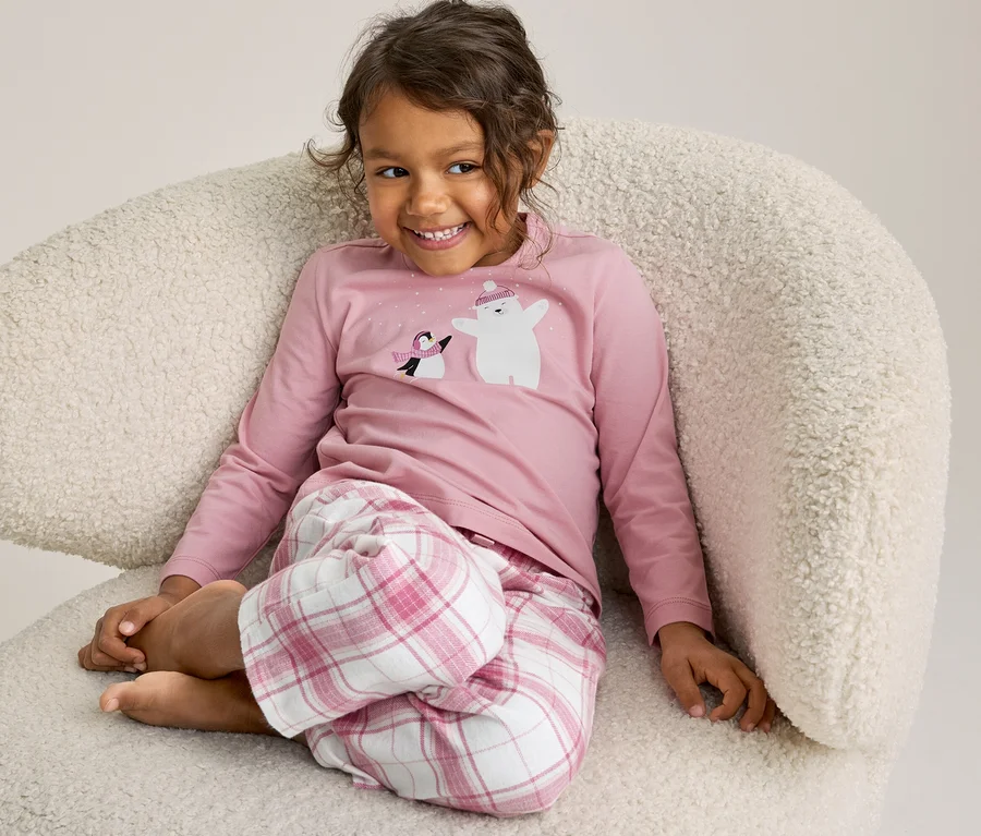 Junges Mädchen mit lockigem Haar sitzt lächelnd auf einem flauschigen weißen Sessel und trägt einen rosa Pyjama mit Eisbär- und Pinguinmotiv.