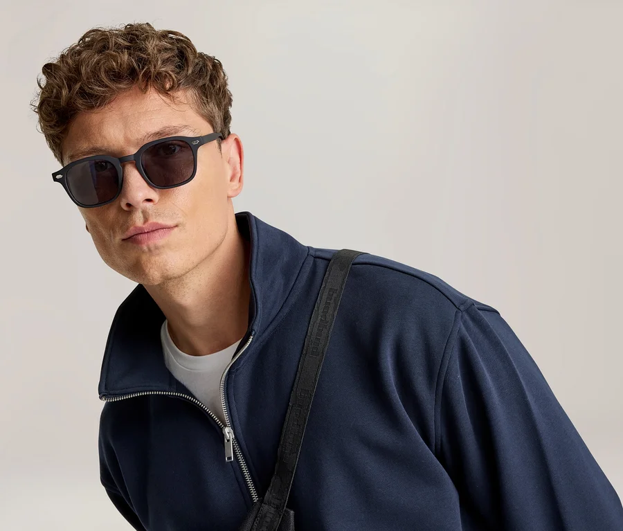 Mann mit lockigem Haar trägt dunkle, eckige Sonnenbrille, eine marineblaue Reißverschlussjacke und ein weißes T-Shirt, blickt in die Kamera.