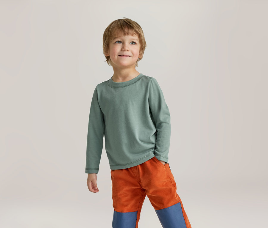 Ein kleiner Junge mit hellbraunen Haaren trägt ein grünes Langarmshirt und orangefarbene Hosen mit blauen Knieflicken. Er steht lächelnd da und blickt nach oben.