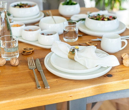 Ein Holztisch ist mit weißen Keramiktellern und Schalen des Mäser Teller-Sets »Taya« (12-tlg) gedeckt, teilweise gefüllt mit Salat und Snacks. Eine weiße Serviette mit Blumen-Serviettenring, Besteck, Wassergläser und dekorative Walnüsse liegen auf dem Tisch.