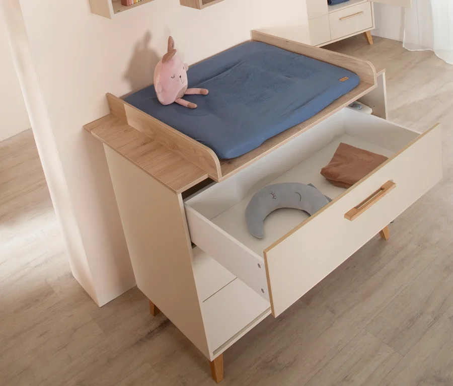 Heller Wickeltisch aus Holz und Weiß mit einer blauen Wickelauflage und einem rosa Stofftier darauf. Eine offene Schublade zeigt ein graues Halbmondkissen und ein braunes Tuch.