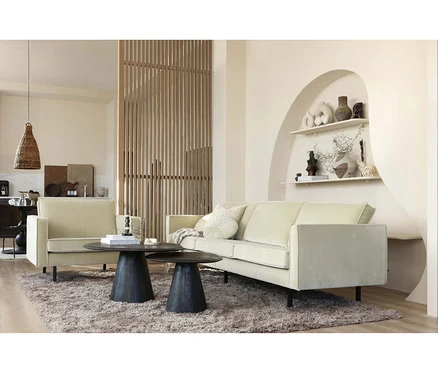 Ein helles Wohnzimmer mit einem beigen Sofa und Sessel, zwei dunklen runden Couchtischen auf einem flauschigen Teppich und einer Holztrennwand. Eine Nische mit Regalen und Dekorationen ist in die Wand eingelassen.