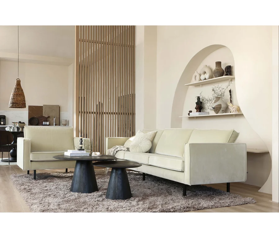 Ein helles Wohnzimmer mit einem beigen Sofa und Sessel, zwei dunklen runden Couchtischen auf einem flauschigen Teppich und einer Holztrennwand. Eine Nische mit Regalen und Dekorationen ist in die Wand eingelassen.