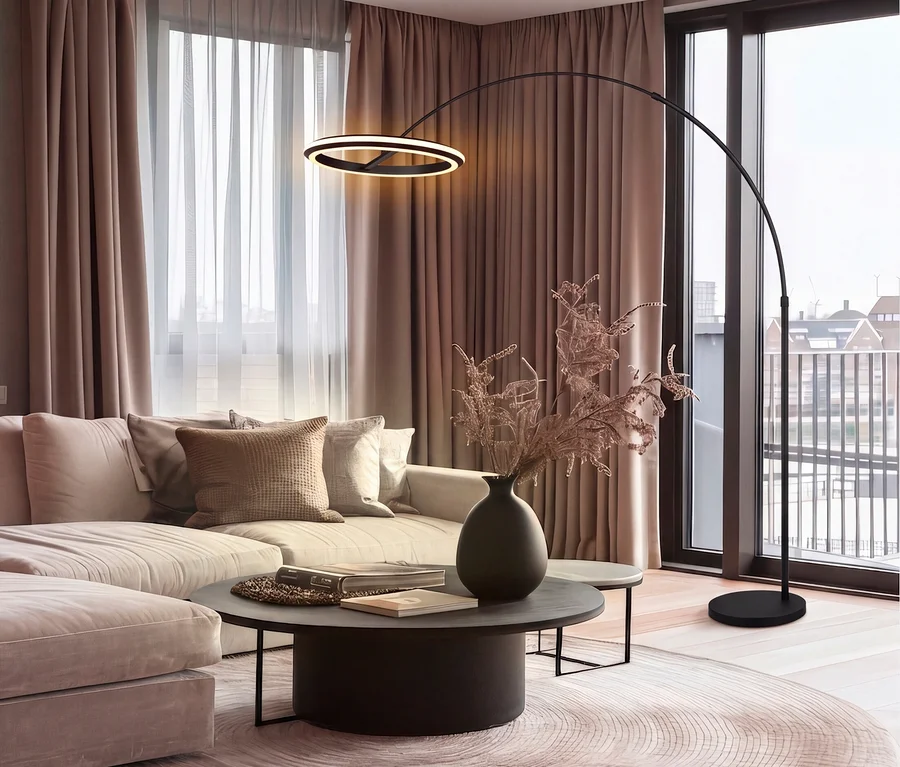 Ein modernes Wohnzimmer mit einem hellbeigen Ecksofa mit Zierkissen, einem dunklen runden Couchtisch mit einer Vase getrockneter Zweige und einer schwarzen Bogenstehlampe mit leuchtendem Ringlicht. Ein runder gemusterter Teppich liegt auf einem Holzfußboden vor großen Fenstern mit taupefarbenen Vorhängen.