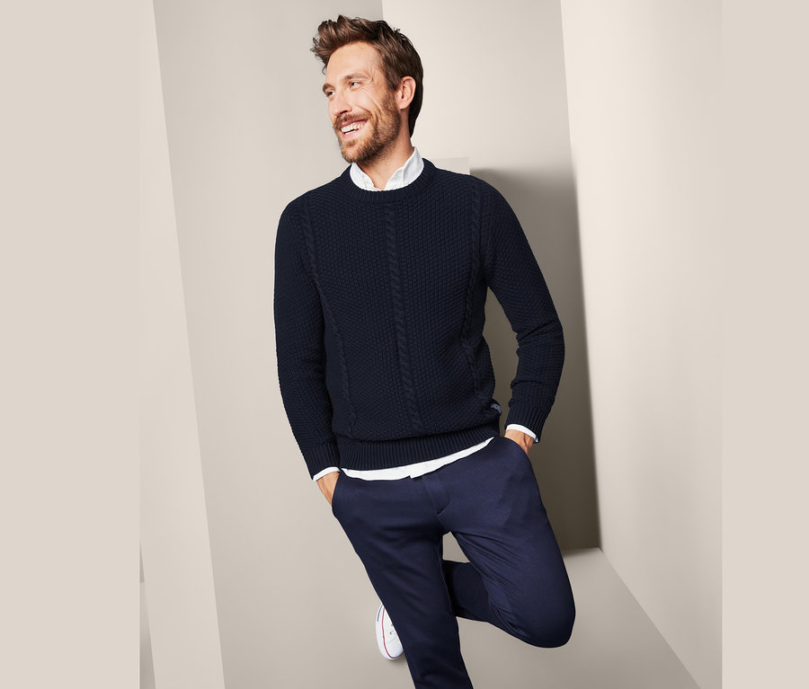 Mann lächelt und lehnt an einer Wand in einem Strickpullover.