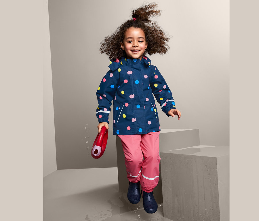 Lächelndes, lockiges Mädchen springt in Thermo-Regenjacke mit bunten Punkten, rosafarbener Hose und blauen Gummistiefeln und hält eine rote Gießkanne.