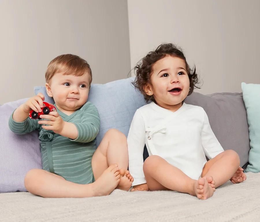 Zwei Babys sitzen auf einem Bett, eines spielt mit einem roten Spielzeugauto, beide tragen 2 Baumwoll-Langarmbodys.