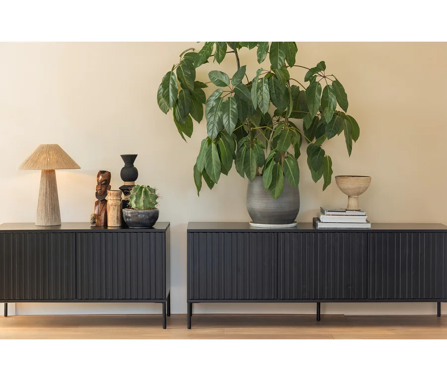 Zwei schwarze Holzsideboards mit geriffelten Türen stehen vor einer hellbeigen Wand auf einem Holzboden. Auf dem linken Sideboard befinden sich eine geflochtene Lampe, Holzfiguren, eine schwarze Vase und ein Kaktus. Das rechte Sideboard trägt eine große Topfpflanze, einen Stapel Bücher und eine Dekoschale.