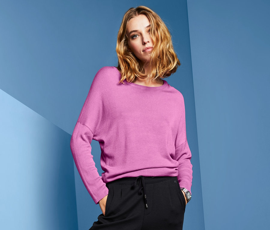 Frau posiert in Feinstrickpullover mit Fledermausärmeln in Pink, schwarzer Hose und silbernen Schuhen.