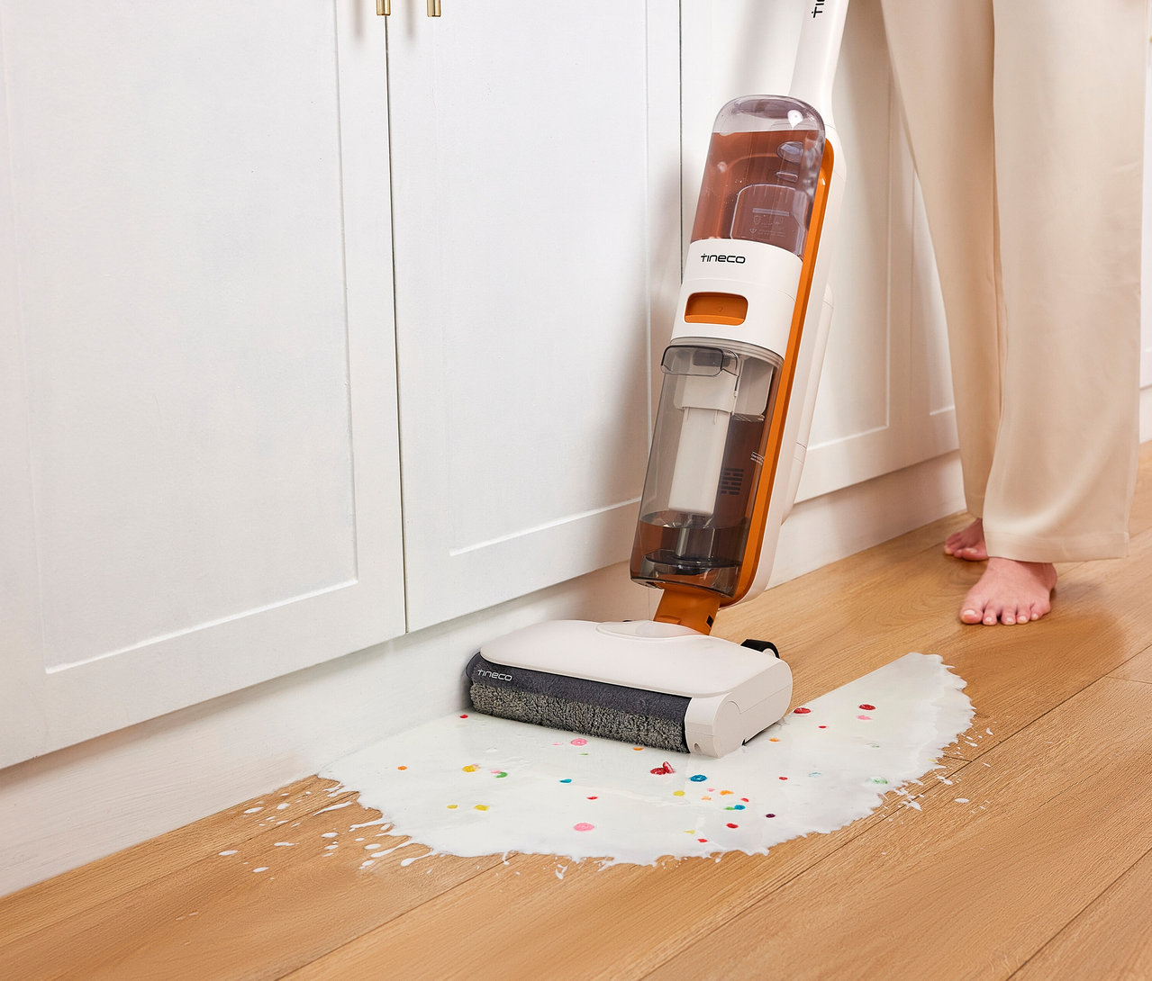 Eine Person mit nackten Füßen reinigt mit dem weißen und orangefarbenen Tineco 2-in-1-Nass-Trockensauger »Y2 Plus« eine verschüttete Flüssigkeit mit bunten Streuseln auf einem Holzboden vor weißen Schränken.