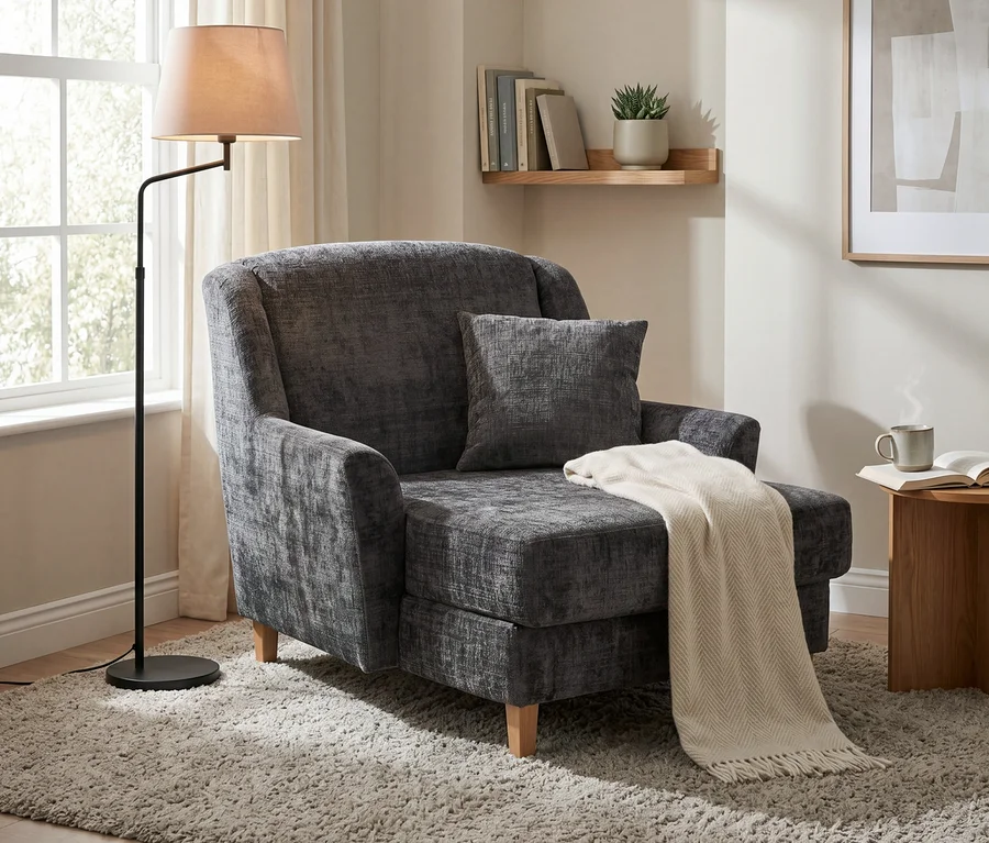 Anthrazitfarbener Big-Sessel „Julia“ mit Chenillebezug und heller Decke auf einem Teppich, daneben eine Stehlampe am Fenster und ein Beistelltisch mit Tasse und Buch.