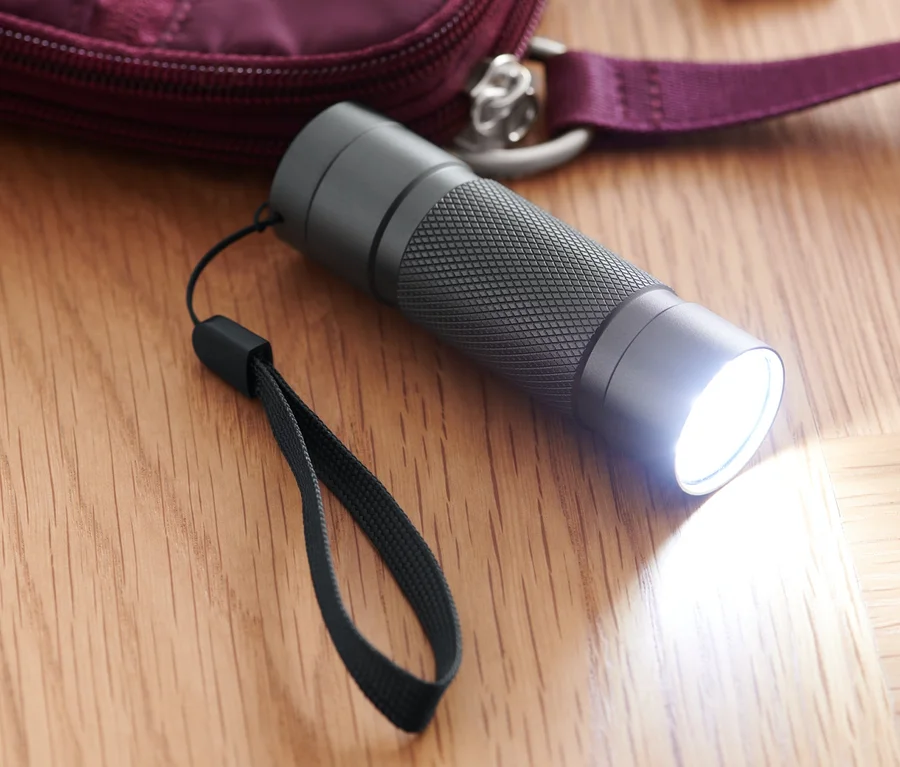 Eine leuchtende LED-Taschenlampe liegt auf einem Holzboden neben einer weinroten Handtasche und Schlüsseln.