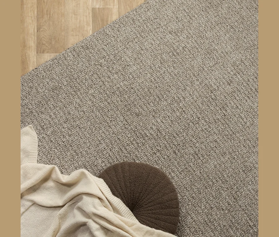 Hellbrauner Teppich bedeckt den Großteil des Bodens, mit einem Holzboden oben links sichtbar. Eine cremefarbene Strickdecke liegt unten links über einem runden, dunkelbraunen Kissen.