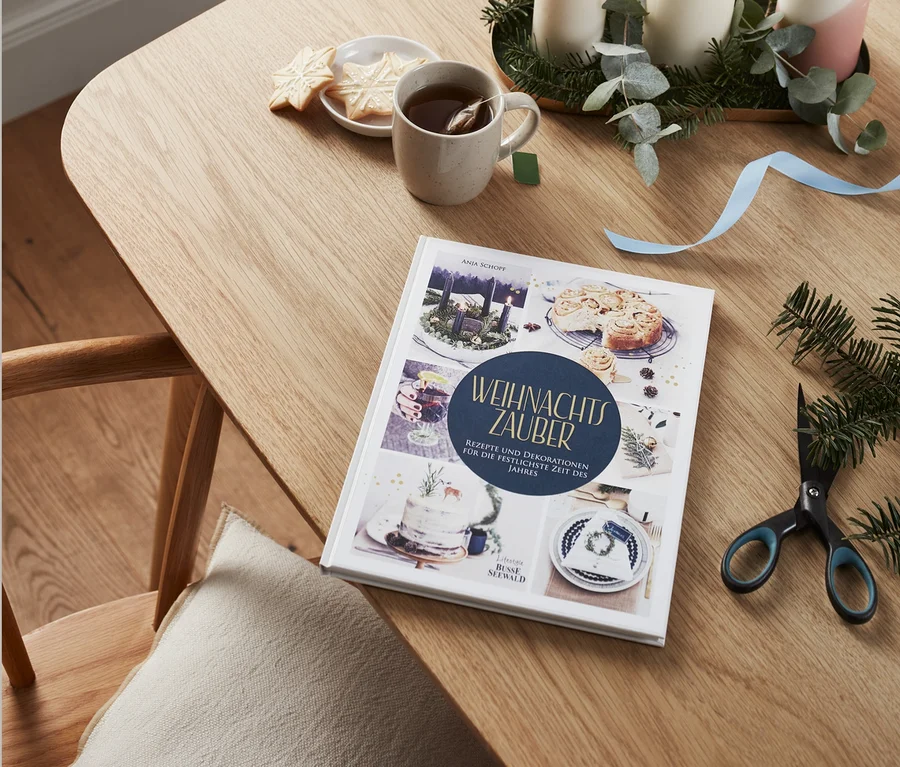 Auf einem Tisch liegen ein Buch "Weihnachtszauber", daneben eine Tasse Tee, ein Teller mit Keksen, Kerzen und eine Schere.