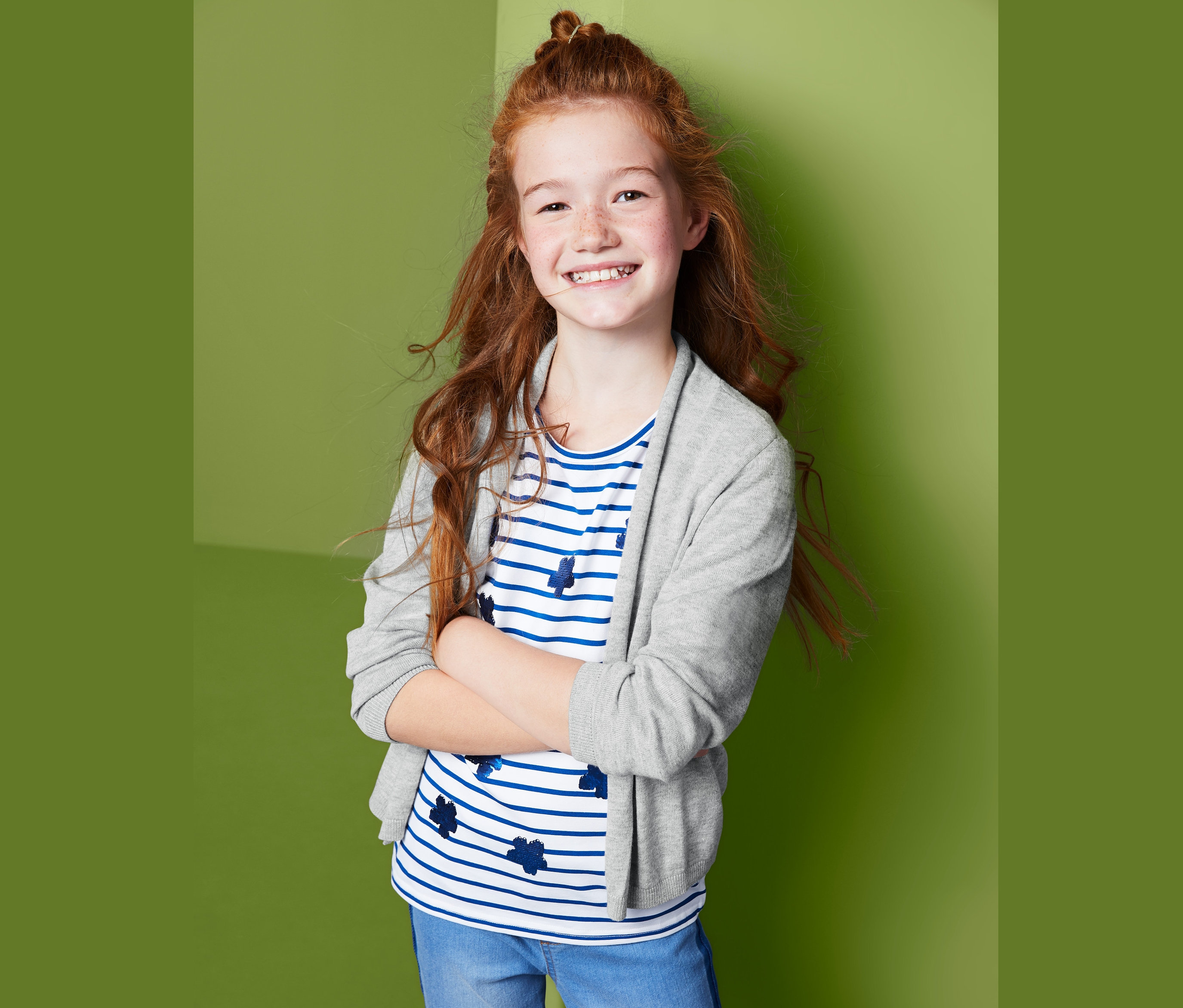 Mädchen mit roten Haaren posiert in offener Strickjacke, blau-weiß gestreiftem T-Shirt und Jeans.