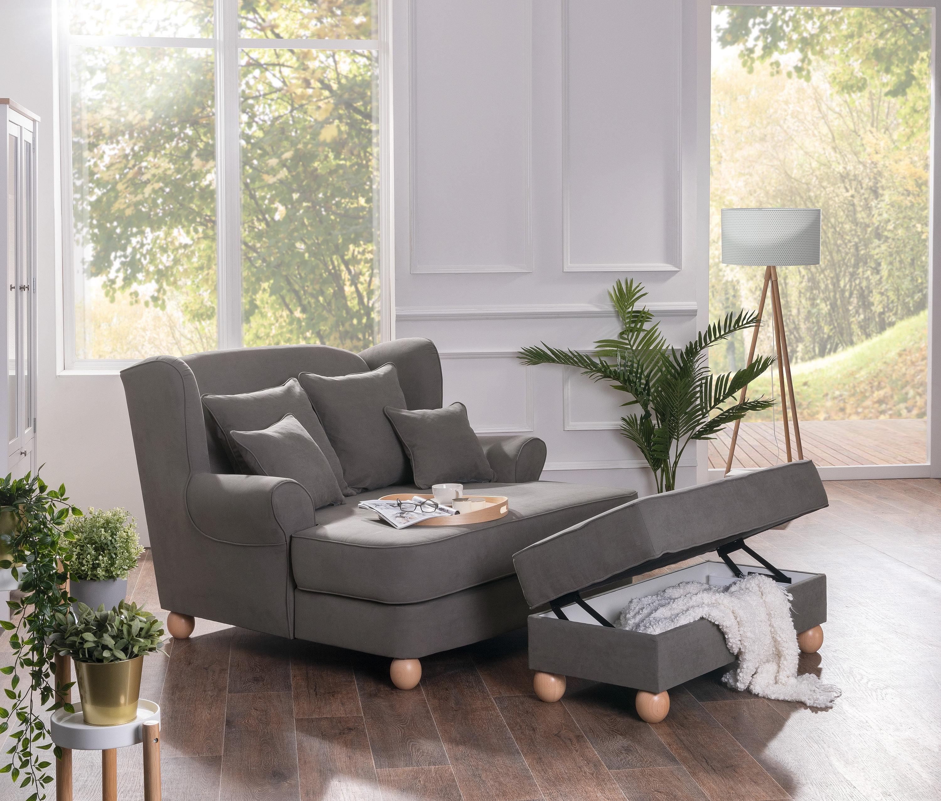 Ein grauer Max Winzer® Polstersessel mit Kissen und aufklappbarer Hockerbank mit Stauraum steht im Wohnzimmer.