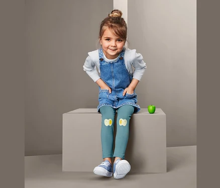 Ein Mädchen sitzt und trägt ein Jeanskleid und 2 Leggings mit Küken-Applikationen.