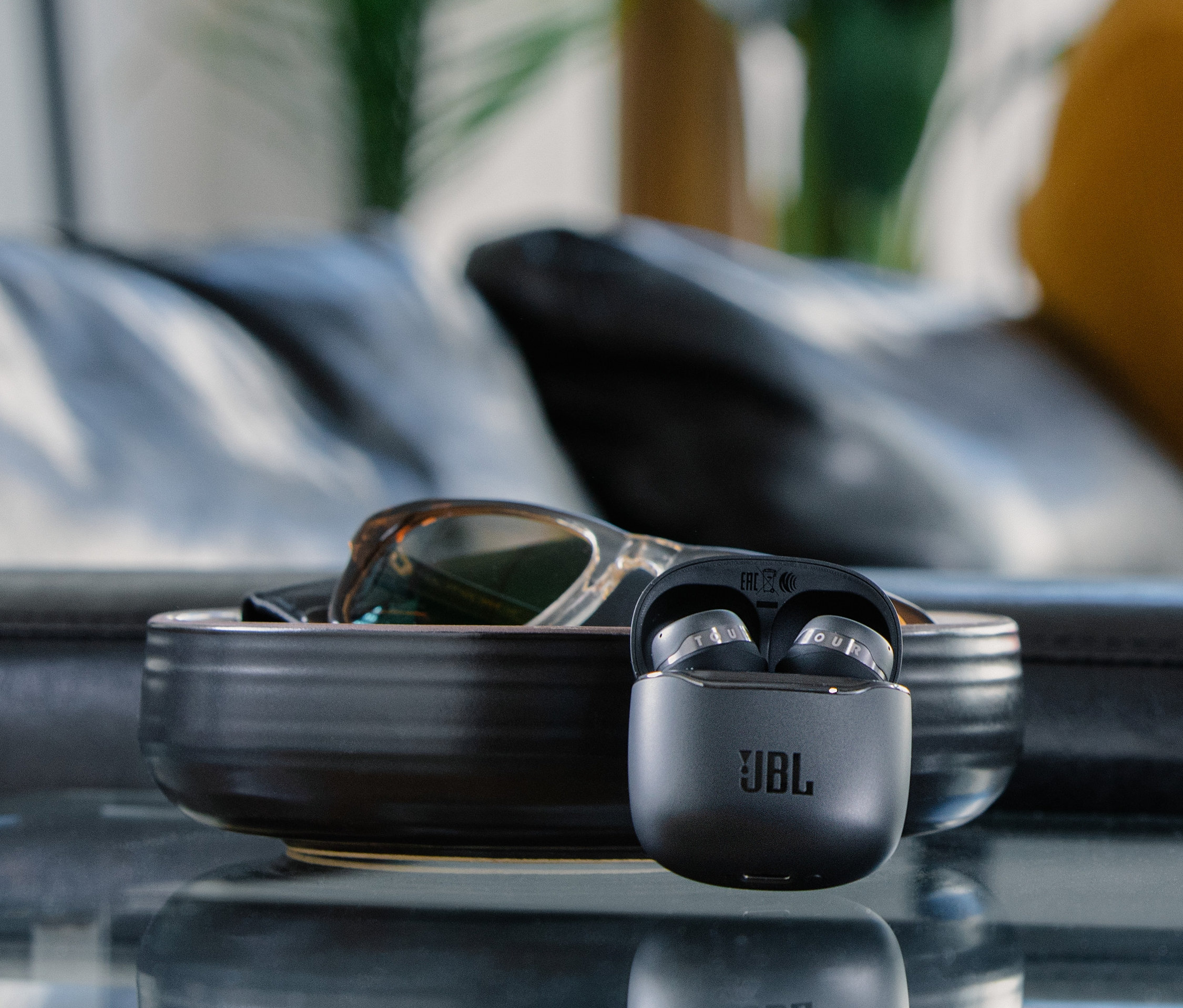 Graue JBL In-Ear Kopfhörer »Tour Pro+ TWS« in Ladebox auf Tisch. Im Hintergrund Brille und Sofa.