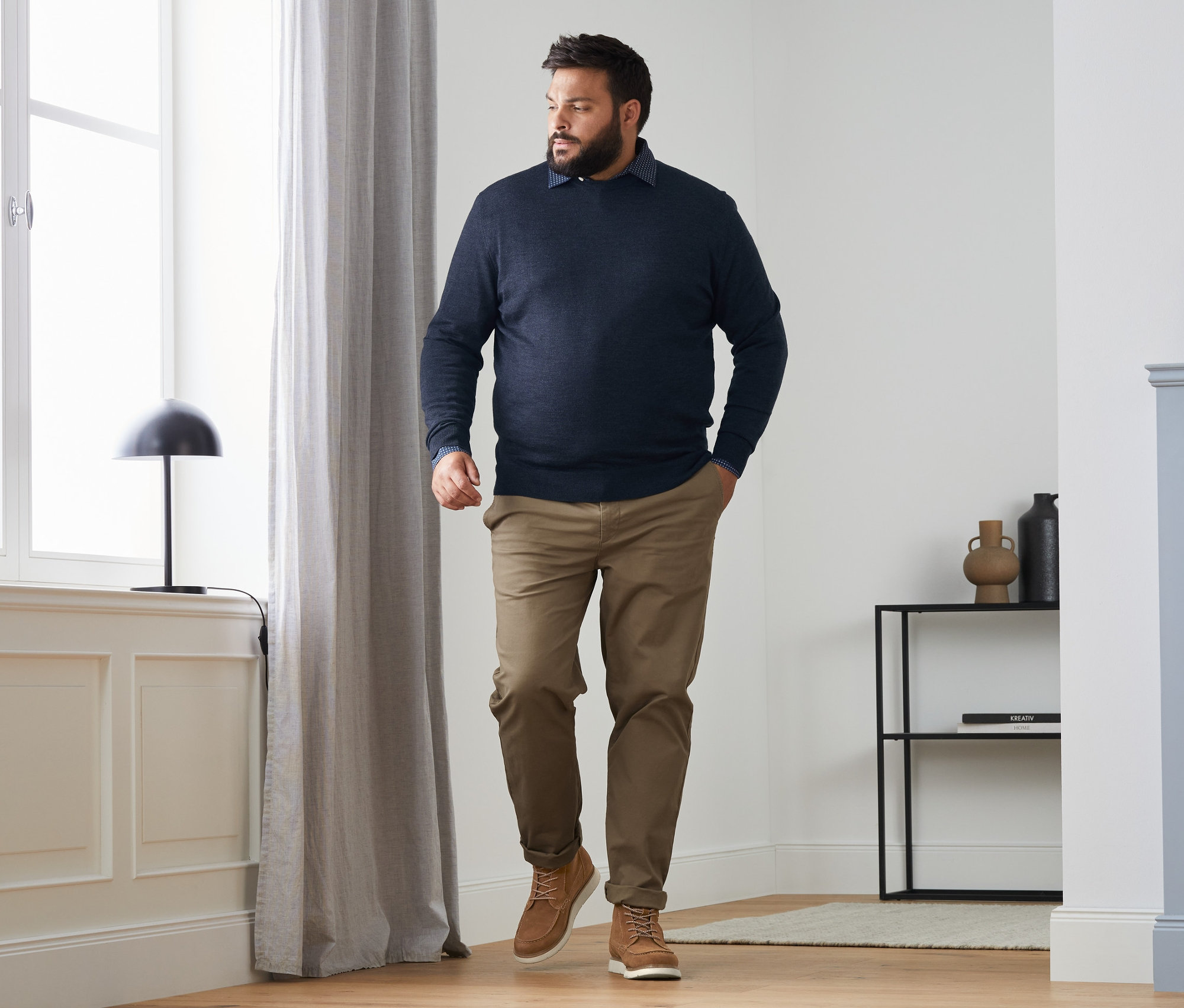 Mann geht in einem dunkelblauen Merino-Pullover mit Rundhalsausschnitt, einer camelfarbenen Chino, einem Hemd mit Comfort Fit und Veloursleder-Boots.