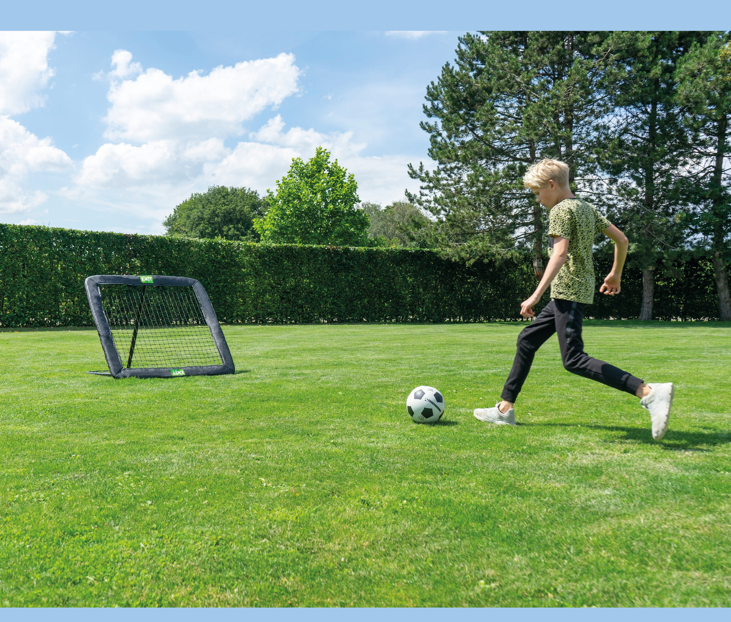 Junge schießt einen Fußball in Richtung EXIT Fußball-Rebounder L auf einer Wiese.