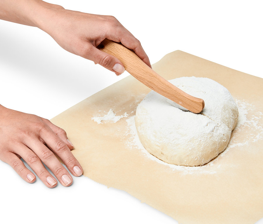 Eine Hand benutzt ein Bäckermesser, um einen Laib Brot auf Backpapier anzuschneiden.