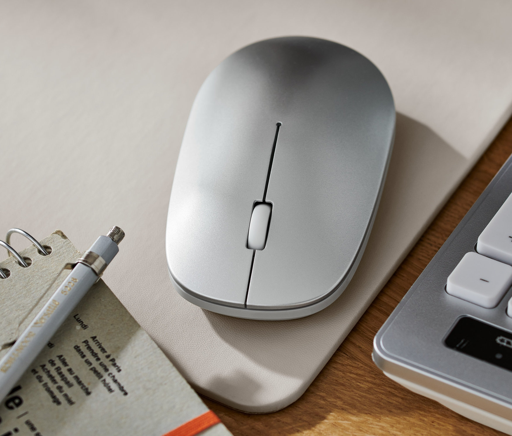 Eine kabellose Maus liegt auf einer Unterlage neben einem Block, einem Stift und einer Tastatur.