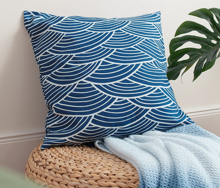 Ein Dekokissen-Bezug mit Wellenprint liegt auf einem Strohhocker mit einer blauen Decke, daneben steht eine Pflanze in einem Strohkorb.