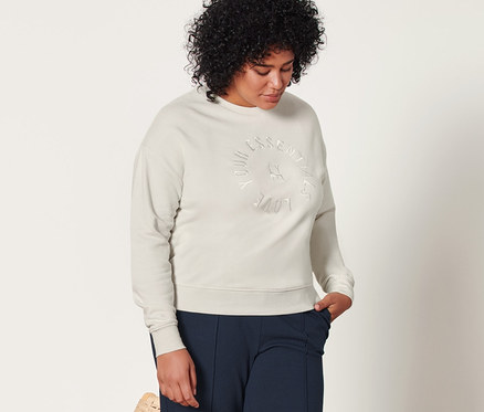 Eine Frau steht neben einem Korbstuhl und trägt eine cremefarbene Sweatshirt und dunkelblaue Joggpants mit Biese.