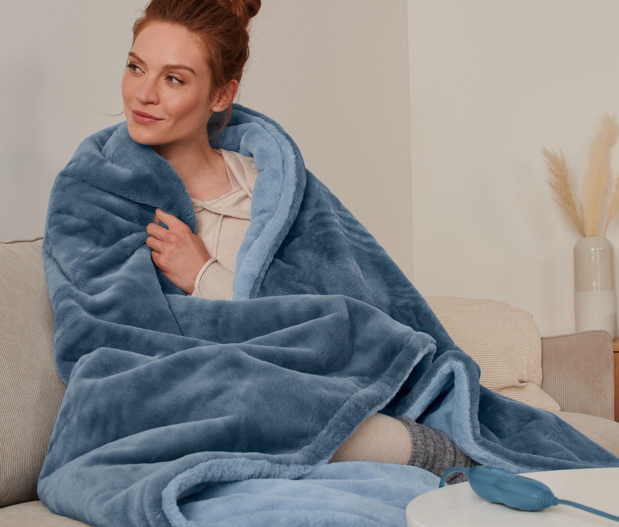 Frau sitzt in eine Heizdecke gehüllt auf einem Sofa.