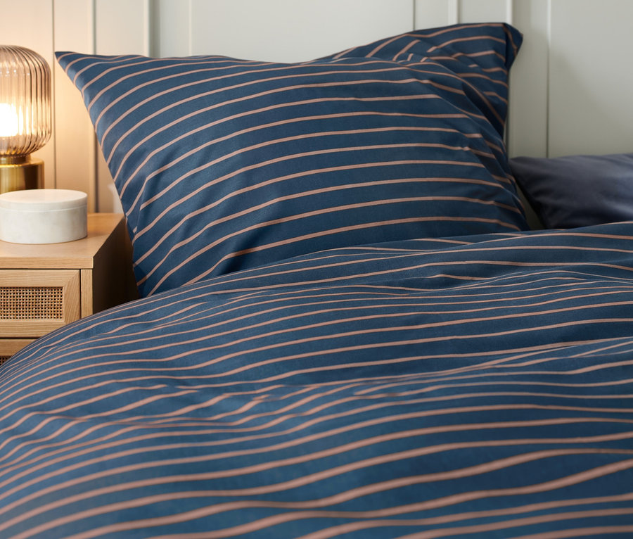 Detailaufnahme eines Schlafzimmers mit Premium-Baumwoll-Bettwäsche mit Streifen auf dem Bett und einem Nachttisch mit Lampe.