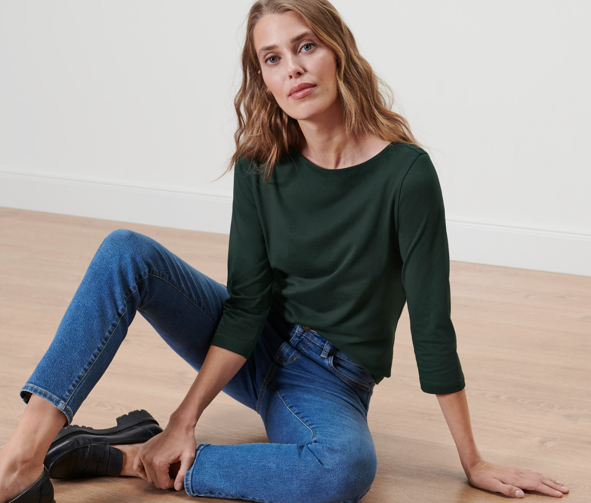 Frau sitzt in Jeans und einem Shirt mit 3/4-Arm auf dem Boden.