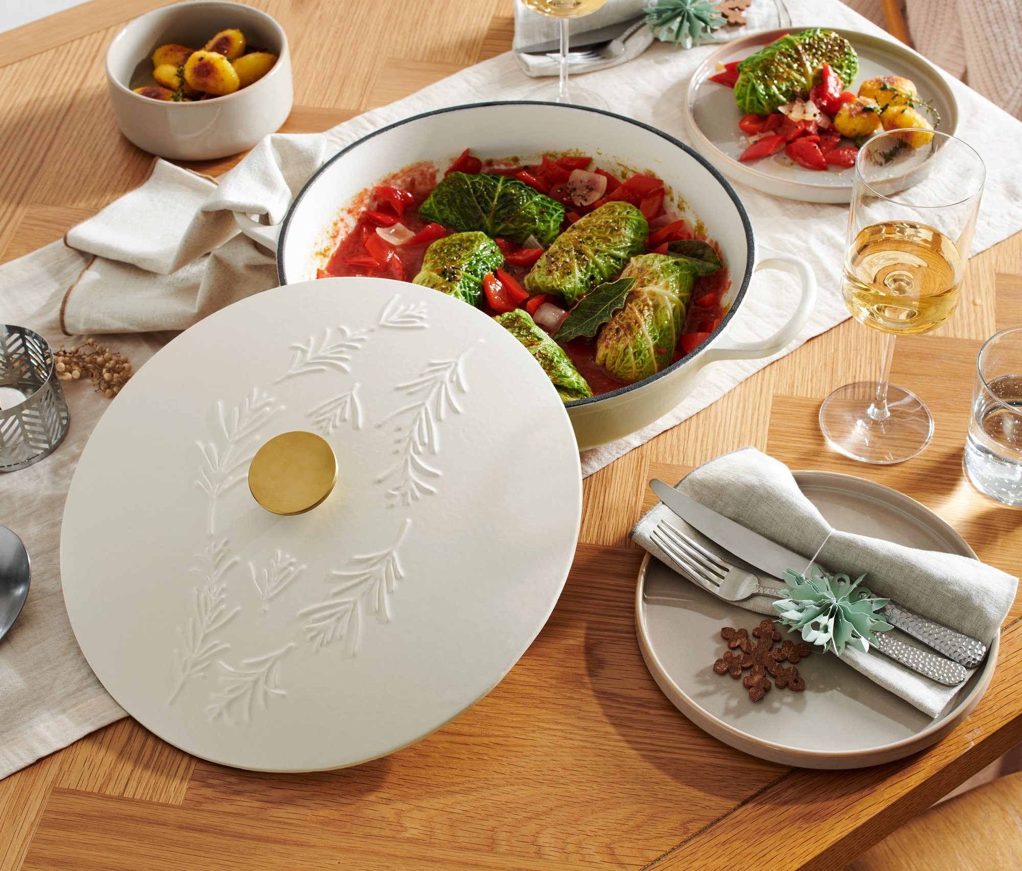 Auf einem Holztisch steht eine Gusseisen-Servierpfanne in Weiß mit einem Deckel, der ein erhabenes Zweigmuster aufweist.